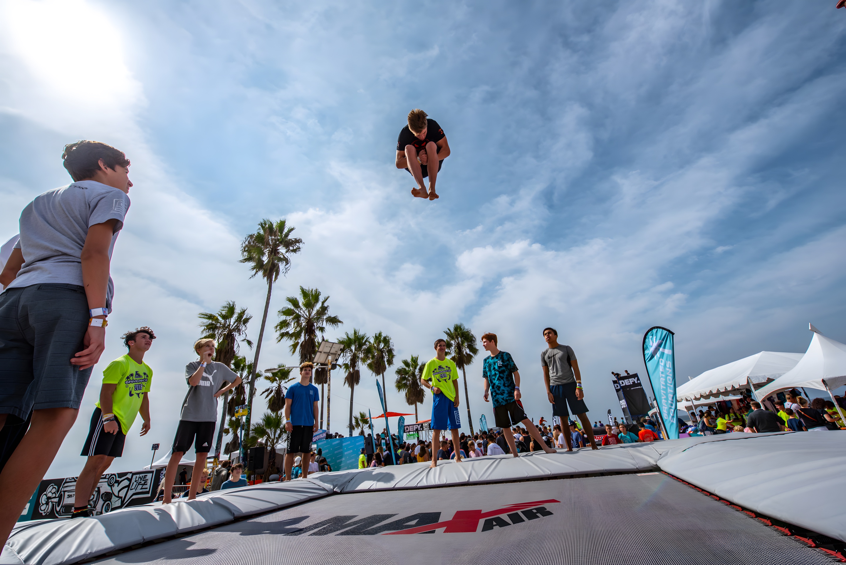 An athlete tucked high in the sky above a MaxAir super trampoline, palm trees and a crowd far below