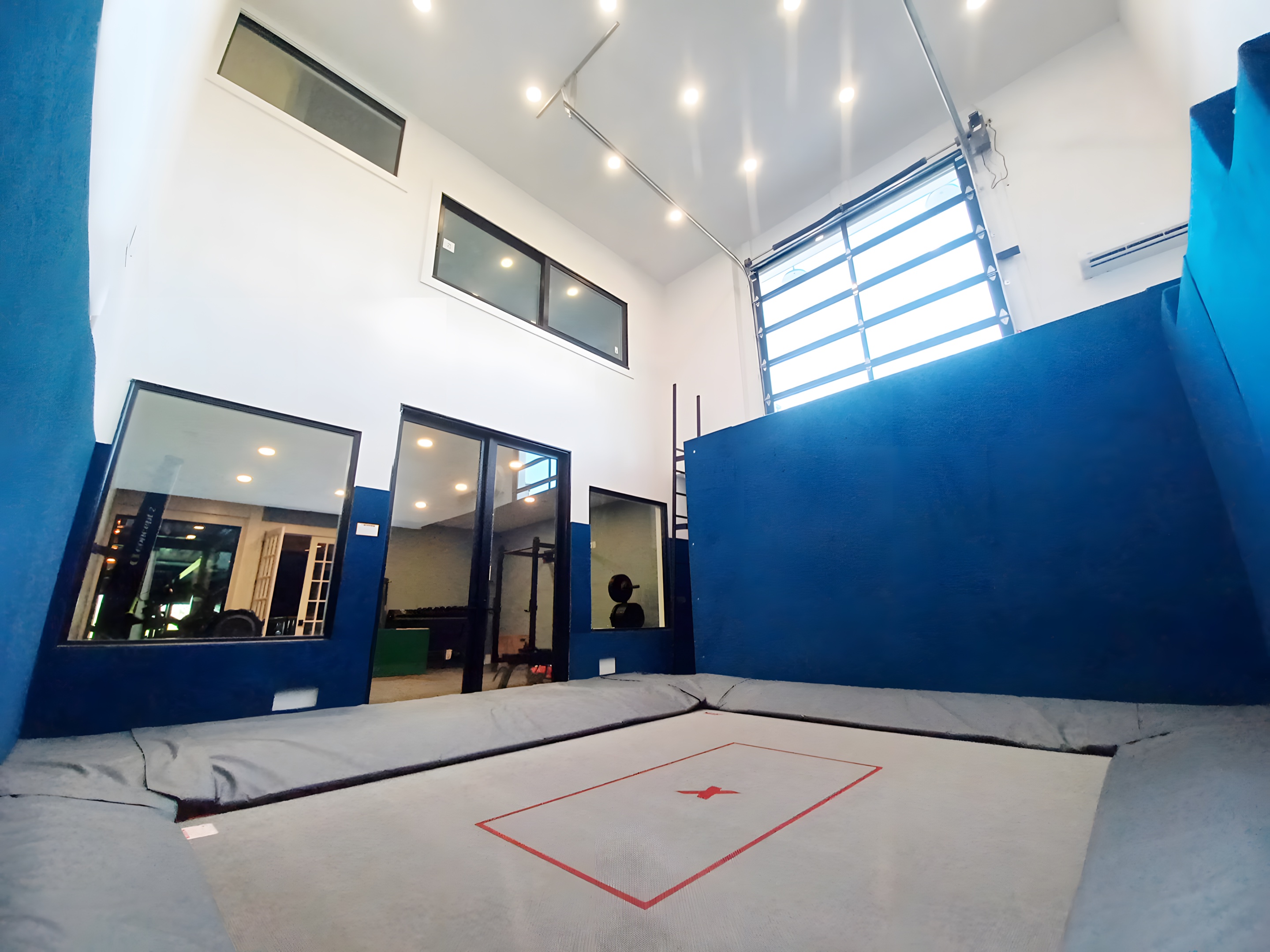 A finished double-height trampoline room with blue padded walls, a sunken MaxAir bed and a glass wall onto the home gym