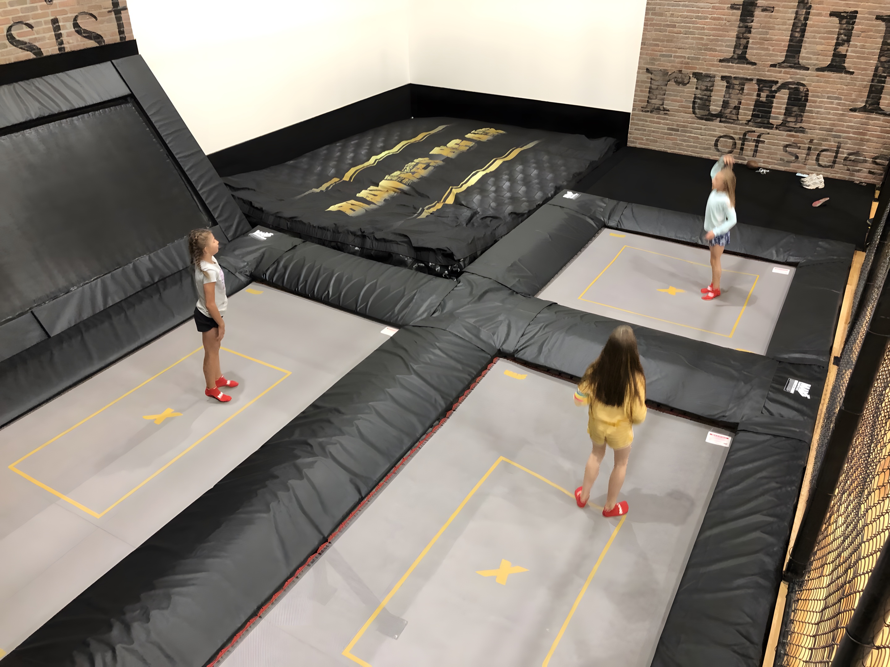 Kids on the sunken beds of a home trampoline room with thick black pads, a trampwall and an air bag
