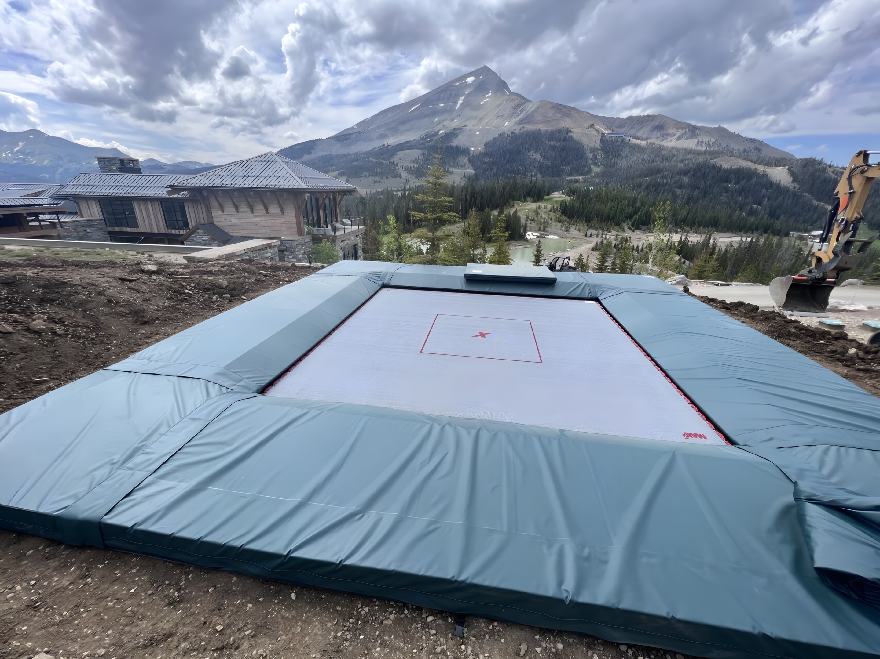 An in-ground super trampoline with thick teal pads set into the ground below a mountain peak